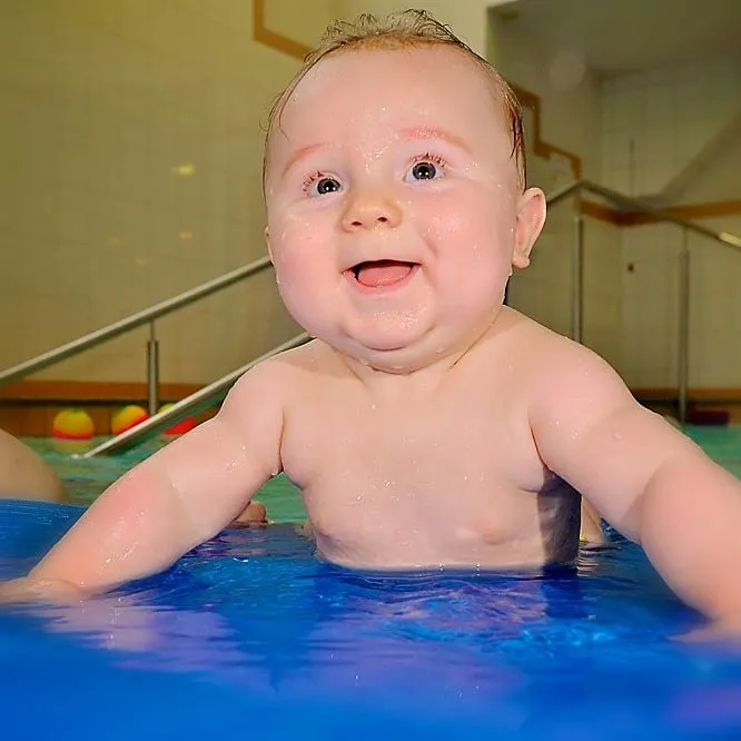Ein Baby stützt sich auf eine blaue Matte im Becken und lacht in die Kamera