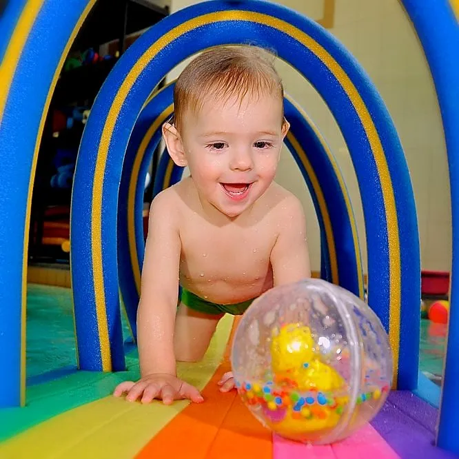 Ein Kleinkind krabbelt über eine bunte Matte unter Bögen aus Poolnudeln hindurch, davor ein Ball