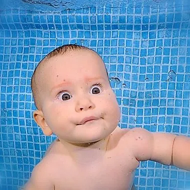 Ein Baby unter Wasser vor einer blau gekachelten Wand, mit offenen Augen