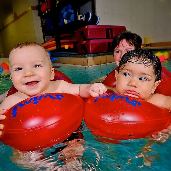 Zwei Babys sitzen nebeneinander in roten Schwimmringen im Becken, dahinter eine erwachsene Person